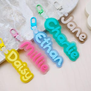 Puede incluir: Una colección de llaveros con nombres coloridos. Cada llavero presenta un nombre diferente: Daisy, Millie, Ethan, Charlie y Jane. Los nombres están en una fuente divertida y están delineados en un color contrastante. Cada llavero tiene un cierre metálico.
