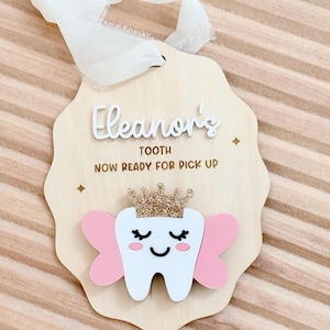 以下が含まれることがあります： 「Eleanor's TOOTH NOW READY FOR PICK UP」の文字が入った木製の歯の妖精の記念品。ピンクの羽と金のグリッタークラウンが付いた笑顔の歯と、吊り下げ用の白いリボンが特徴です。