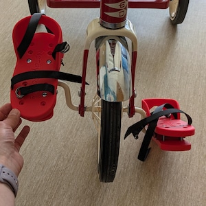 以下が含まれることがあります： 黒色の車輪と黒いストラップが付いた赤と白の三輪車。三輪車は赤い座席とクロム製のハンドルバーが付いています。三輪車は黒いストラップが付いた赤いフットレストが2つあり、フットレストには「Nerdy」という文字が書かれています。