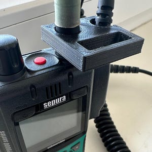 Könnte beinhalten: Ein schwarzes und grünes Zweiwegefunkgerät mit einem schwarzen Bildschirm und dem Wort "sepura" darauf gedruckt. Das Funkgerät hat einen roten Knopf und ein schwarzes Spiralkabel, das daran befestigt ist. Ein schwarzes 3D-gedrucktes Zubehörteil ist oben am Funkgerät befestigt.