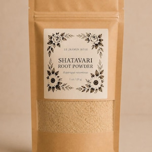 May include: A brown paper bag with a clear window displaying a tan powder. The bag has a white label with the text "LE JASMIN ROSE" and "SHATAVARI ROOT POWDER" in a floral design. The label also states "1 oz / 28 g".