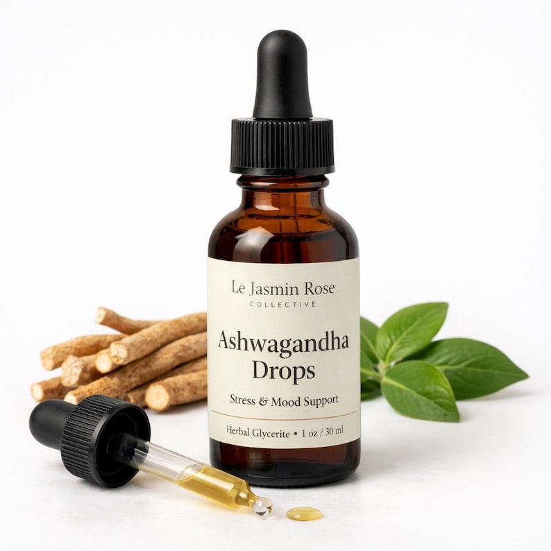 May include: A brown glass bottle of Ashwagandha Drops with a black dropper, next to a pile of brown roots and green leaves. The label reads "Le Jasmin Rose Collective" and "Stress & Mood Support". The bottle contains 1 oz / 30 ml of herbal glycerite.