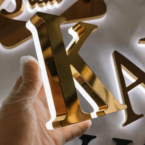 Peut inclure: Une lettre "K" dorée et brillante est tenue dans une main, avec une lumière blanche soulignant les bords. D'autres lettres dorées sont visibles en arrière-plan. Les lettres semblent faire partie d'une enseigne ou d'un présentoir décoratif.