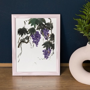 May include: A framed watercolor painting of purple grapes and green leaves. The artwork is in a light pink frame and is set against a dark blue wall. A white vase with a circular opening and a fern are to the right.