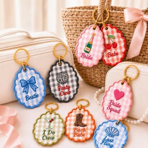May include: A collection of scalloped, gingham-patterned keychains with gold rings. Designs include a blue bow labeled Bestie, a gray disco ball labeled Party Crew, a green martini labeled I Do Crew, an orange cowboy boot labeled Bach Babe, and pink items.