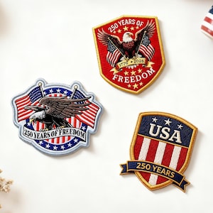 May include: Three embroidered patches with patriotic themes. One patch features an eagle with the text "250 YEARS OF FREEDOM". Another patch displays "USA" and "250 YEARS". A small cup with a star-spangled design is also visible.