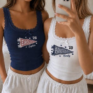 May include: A navy blue ribbed vest top with lace trim and a red gingham pennant graphic reading 'EAGLES '26 Sophia' and a white ribbed vest top with lace trim and a blue gingham pennant graphic reading 'PANTHERS '10 Emily', both paired with white shorts.