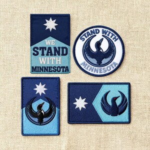 May include: Four embroidered patches with a navy blue and light blue color scheme. The patches feature a white star, a bird graphic, and the text "WE STAND WITH MINNESOTA" and "STAND WITH MINNESOTA".