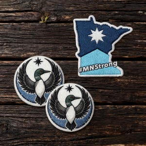 May include: Three embroidered patches on a wooden surface. Two patches feature a bird with outstretched wings, a star, and a blue and white border. The third patch is in the shape of a state with a star and the text "#MNStrong".