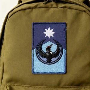 May include: Olive green backpack with a rectangular patch featuring a black bird design, a white star, and blue accents. The patch is stitched onto the backpack.