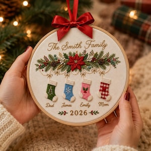 May include: A round wooden embroidery hoop featuring a cream fabric background with a red satin bow at the top. The design includes embroidered pine branches, a red poinsettia, and four stockings labeled Dad, James, Emily, and Mom. Text reads The Smith Family and 2026.