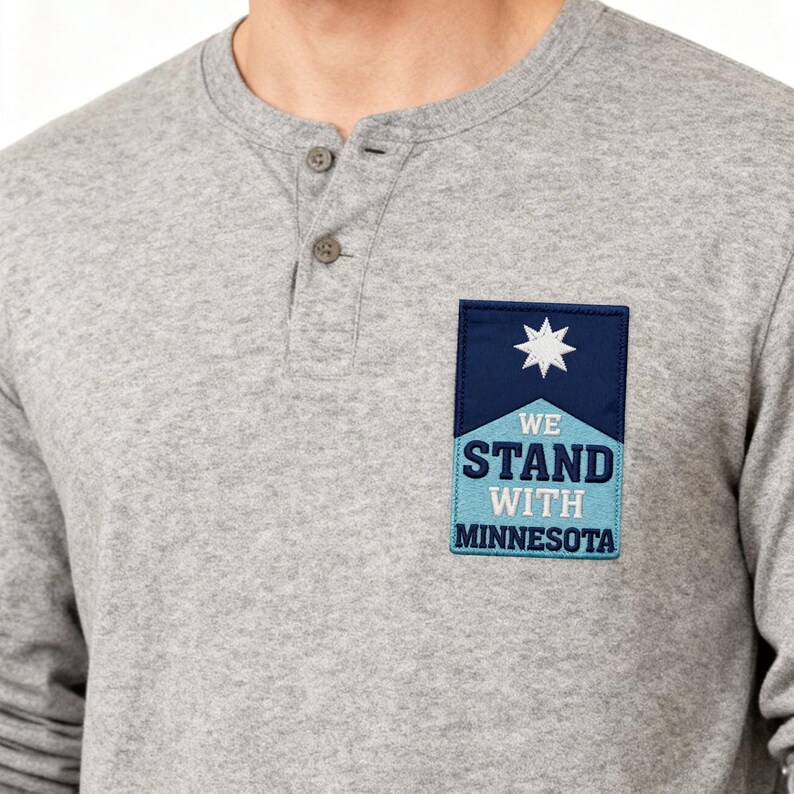 May include: A gray long-sleeve Henley shirt with a three-button placket. A patch on the chest reads "WE STAND WITH MINNESOTA" with a star design. The shirt is made of a soft, comfortable material.