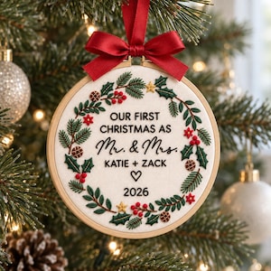 May include: A round wooden embroidery hoop ornament featuring a white fabric center with a hand-stitched wreath of green pine needles, red berries, and small gold stars. The text reads: OUR FIRST CHRISTMAS AS Mr. & Mrs. KATIE + ZACK 2026.