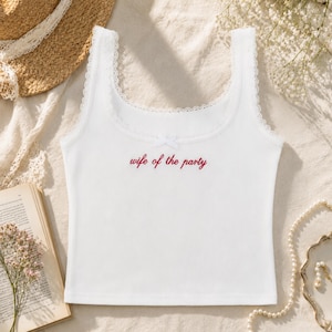 May include: White tank top with a lace trim and a small bow detail. The text "wife of the party" is embroidered in red script across the front. The top is made of a soft, lightweight material.