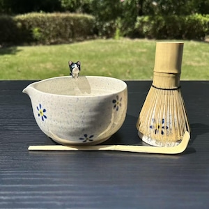 Puede incluir: Un cuenco de matcha de cerámica con detalles florales azules, un batidor de bambú y una cuchara de bambú. Una pequeña figura de gato de cerámica está colocada en el borde del cuenco. El conjunto está sobre una superficie oscura.
