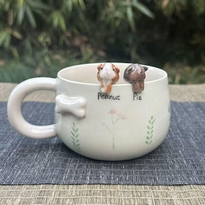 May include: Cream-colored ceramic mug with a bone-shaped handle and two small guinea pig figurines perched on the rim. The mug is decorated with painted flowers and leaves, and the names "Peanut" and "Pia" are written below the figurines.