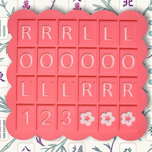 May include: A coral-colored, scalloped-edge mold for making decorative tiles. The mold has a grid of rectangular spaces with the letters R, L, and O, numbers 1, 2, and 3, and flower designs. The background features a floral and Mahjong tile pattern.