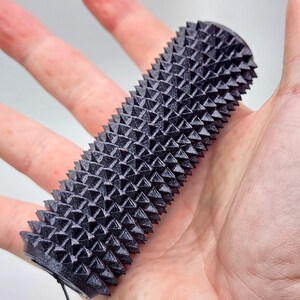 May include: A black massage tool with a cylindrical shape and a textured surface covered in pyramid-shaped spikes. The tool is held in an open hand. The massage tool is designed for muscle relief and is approximately 10 cm in length.