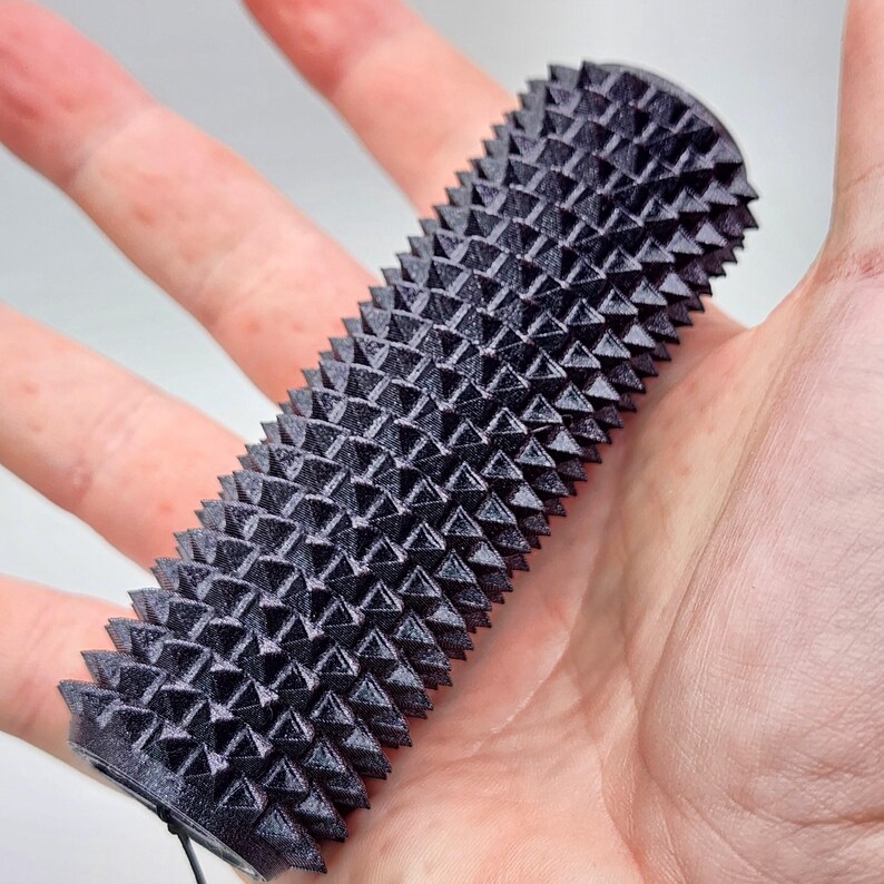 May include: A black massage tool with a cylindrical shape and a textured surface covered in pyramid-shaped spikes. The tool is held in an open hand. The massage tool is designed for muscle relief and is approximately 10 cm in length.