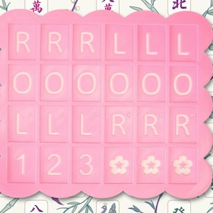 May include: A pink, scalloped-edged tray with a grid of lettered and numbered tiles. The tiles spell out "RRR LLL OOO LLL RRR 123" and feature three flower designs. The background has a floral and Asian-inspired pattern.