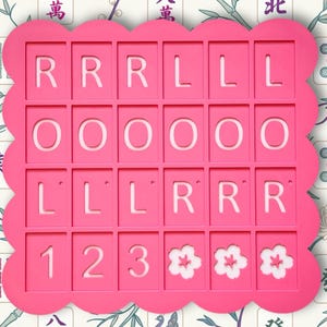 May include: A bright pink, scalloped-edge game board with cut-out squares. The squares contain the letters R, O, and L, along with the numbers 1, 2, and 3, and three white flower designs. The background features a floral and bamboo pattern.