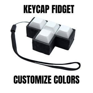 May include: A black keycap fidget with white keycaps, connected to a black lanyard. The text "KEYCAP FIDGET" and "CUSTOMIZE COLORS" are displayed in bold, black font.