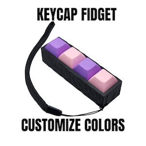 May include: A black keycap fidget with four square keycaps in pink and purple. The keycaps are in a black rectangular container with a black wrist strap. Text at the top says "KEYCAP FIDGET" and at the bottom "CUSTOMIZE COLORS".