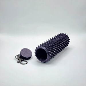May include: A dark purple, cylindrical container with a spiked exterior, next to a matching circular lid attached to a keychain. The container is open, revealing a hollow interior. The design features a series of evenly spaced, sharp spikes.