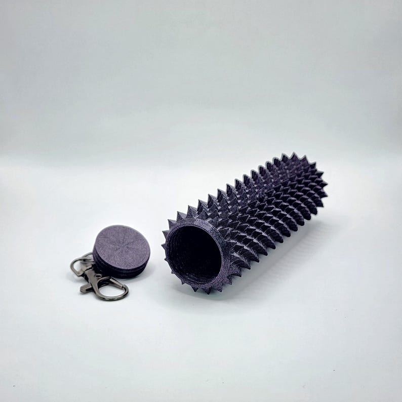 May include: A dark purple, cylindrical container with a spiked exterior, next to a matching circular lid attached to a keychain. The container is open, revealing a hollow interior. The design features a series of evenly spaced, sharp spikes.