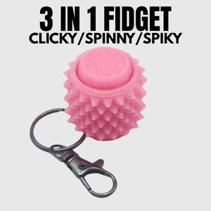 Pode incluir: Um brinquedo fidget rosa 3 em 1 com um design espinhoso, um topo clicável e um porta-chaves. O texto "3 IN 1 FIDGET CLICKY/SPINNY/SPIKY" é exibido acima do brinquedo.