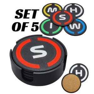 May include: A set of five black coasters with a holder. Each coaster has a black base with a colored ring and a white letter. The holder and coasters have the text "SET OF 5". One coaster has a cork base.