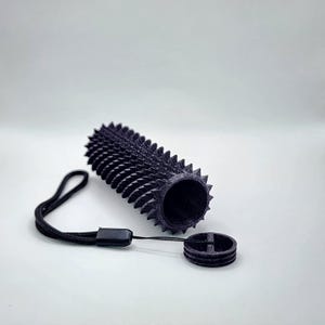 May include: A black, cylindrical object with a textured, spiky exterior, next to a matching circular piece and a black lanyard. The object appears 3D-printed. The background is plain white.
