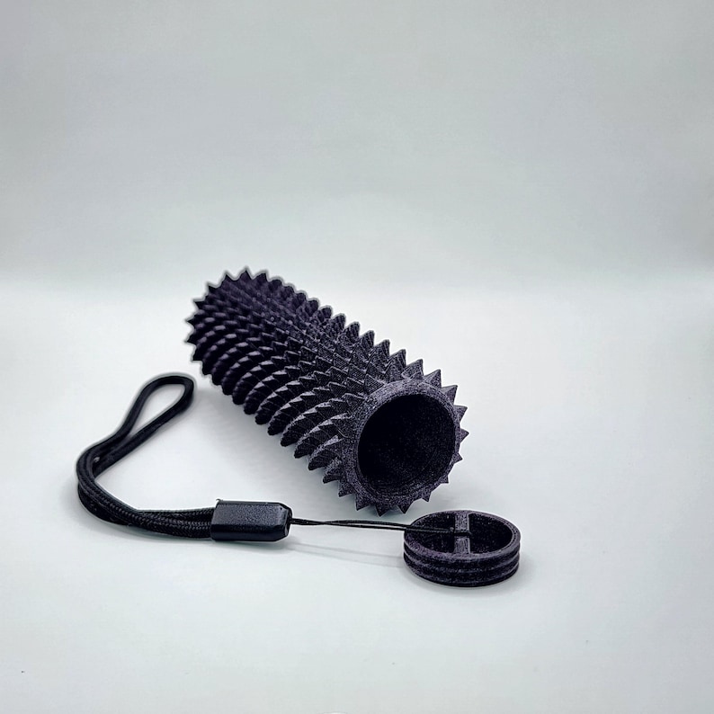 May include: A black, cylindrical object with a textured, spiky exterior, next to a matching circular piece and a black lanyard. The object appears 3D-printed. The background is plain white.