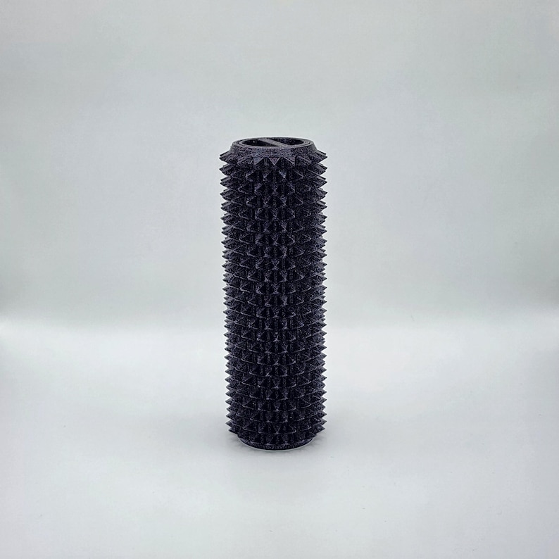 May include: A dark purple, cylindrical object with a textured surface of evenly spaced, conical spikes. The top rim has a decorative, ridged design. The object is set against a plain white background.