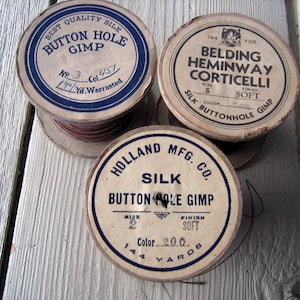 May include: Three spools of brown thread with white labels. The labels have blue text and say "Best Quality Silk Button Hole Gimp", "Belding Heminway Corticelli Silk Buttonhole Gimp", and "Holland Mfg. Co. Silk Button Hole Gimp".
