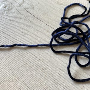 May include: Close-up of navy blue cord, looped and coiled on a light wood surface. The cord has a soft, textured appearance, with variations in the blue hue. The cord is likely used for crafting or decorative purposes.