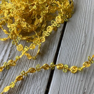 May include: Close-up of a spool of yellow floral trim. The trim is made of a twisted cord with small, delicate rose-shaped embellishments. The trim is on a weathered, white-painted wooden surface.