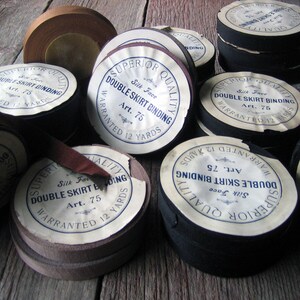 May include: A collection of spools of black and brown double skirt binding with white labels that read "Superior Quality Silk Face Double Skirt Binding Art. 75 Warranted 12 Yards".