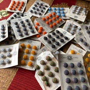 May include: A collection of vintage buttons in various colors, including orange, gray, blue, yellow, and green, arranged on aged paper cards. The buttons feature swirl and dot designs, offering a range of styles for sewing and crafting projects.