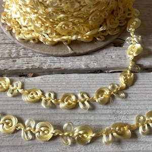 May include: A spool of yellow decorative trim with a floral design. The trim is made of a soft, looped material and is arranged in a continuous pattern. The trim is on a wooden surface.