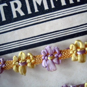 May include: A close-up of a trim with alternating purple and yellow flowers. The trim is made of a woven fabric with a satin finish.