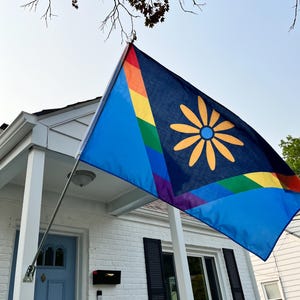 May include: A blue flag with a rainbow stripe and a yellow flower design, hanging from a white porch. The flag is displayed in front of a white brick house with a blue door and black shutters.