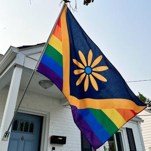 Puede incluir: Una bandera azul marino con un gran diseño de margarita amarilla y una franja arcoíris. La bandera cuelga de una casa blanca con una puerta azul.