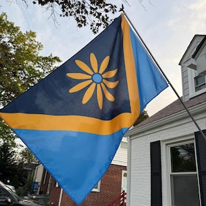 May include: A flag with a blue and yellow design, featuring a large yellow flower in the upper left corner. The flag is mounted on a pole and is displayed outdoors near a white house with black shutters.