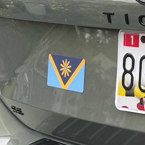 May include: A rectangular car magnet with a floral design. The magnet features a blue and yellow color scheme with a stylized flower in the center. The magnet is affixed to the rear of a gray vehicle.