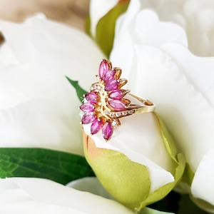 May include: A gold ring featuring a cluster of pink gemstones in a floral design. The ring has a band with small, clear gemstones. The ring is set against a backdrop of white flowers and green leaves.