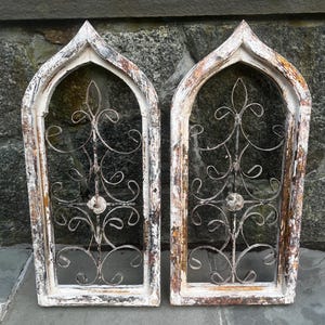 May include: Two arched wall decor pieces featuring weathered white and brown wooden frames with pointed tops. Each frame contains a dark metal insert with symmetrical scrollwork patterns and a central circular medallion detail.