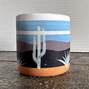 May include: Cylindrical ceramic pot with a hand-painted desert landscape design. The pot features a white cactus, blue sky, and orange base. The design includes a black and brown mountain range and white accents.