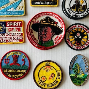 May include: A collection of vintage Boy Scout patches in various shapes and colors. The patches feature text and graphics related to camping, scouting events, and locations. Some patches include text like "Camporee," "Jamboree," and "Scout-O-Rama."