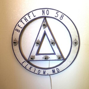 May include: A circular white and purple wall-mounted sign featuring a central triangle and seven light bulb sockets. The text BETHFL NO 58 and ELKTON, MD is printed in purple along the circular frame. A white power cord with a switch hangs below.